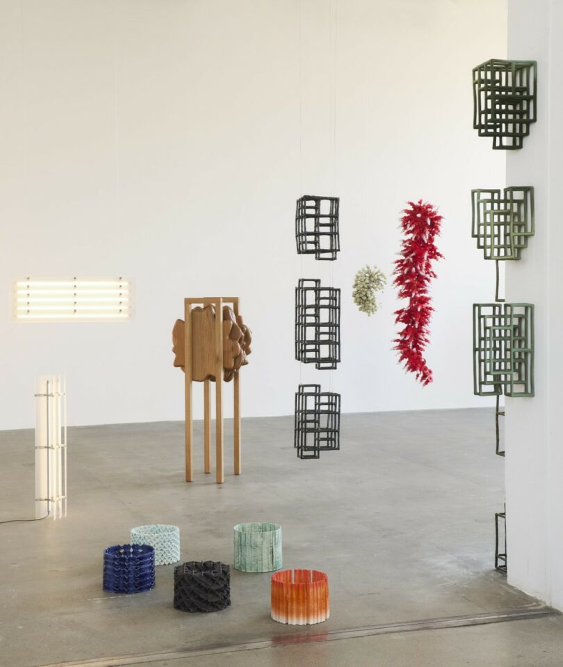 A variety of contemporary furniture and sculptural pieces are arranged in a minimalist gallery with white walls and a concrete floor