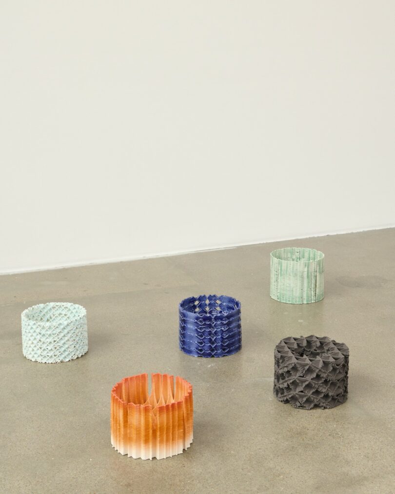 Five cylindrical, textured objects in various colors (blue, orange, green, white, gray) are arranged on a polished concrete floor against a plain white wall