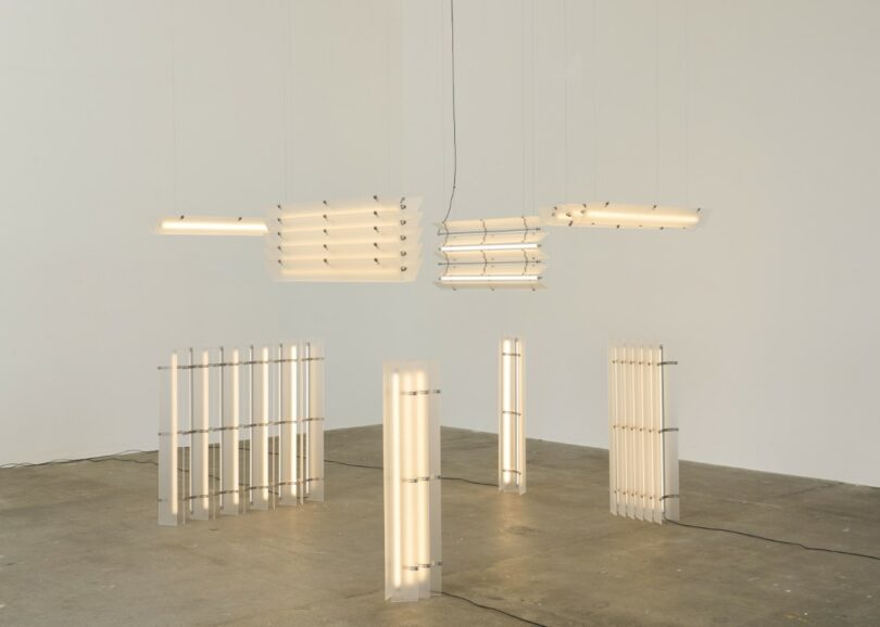 Several rectangular, tube-like light fixtures are suspended from the ceiling and standing on the concrete floor in a minimalist gallery space