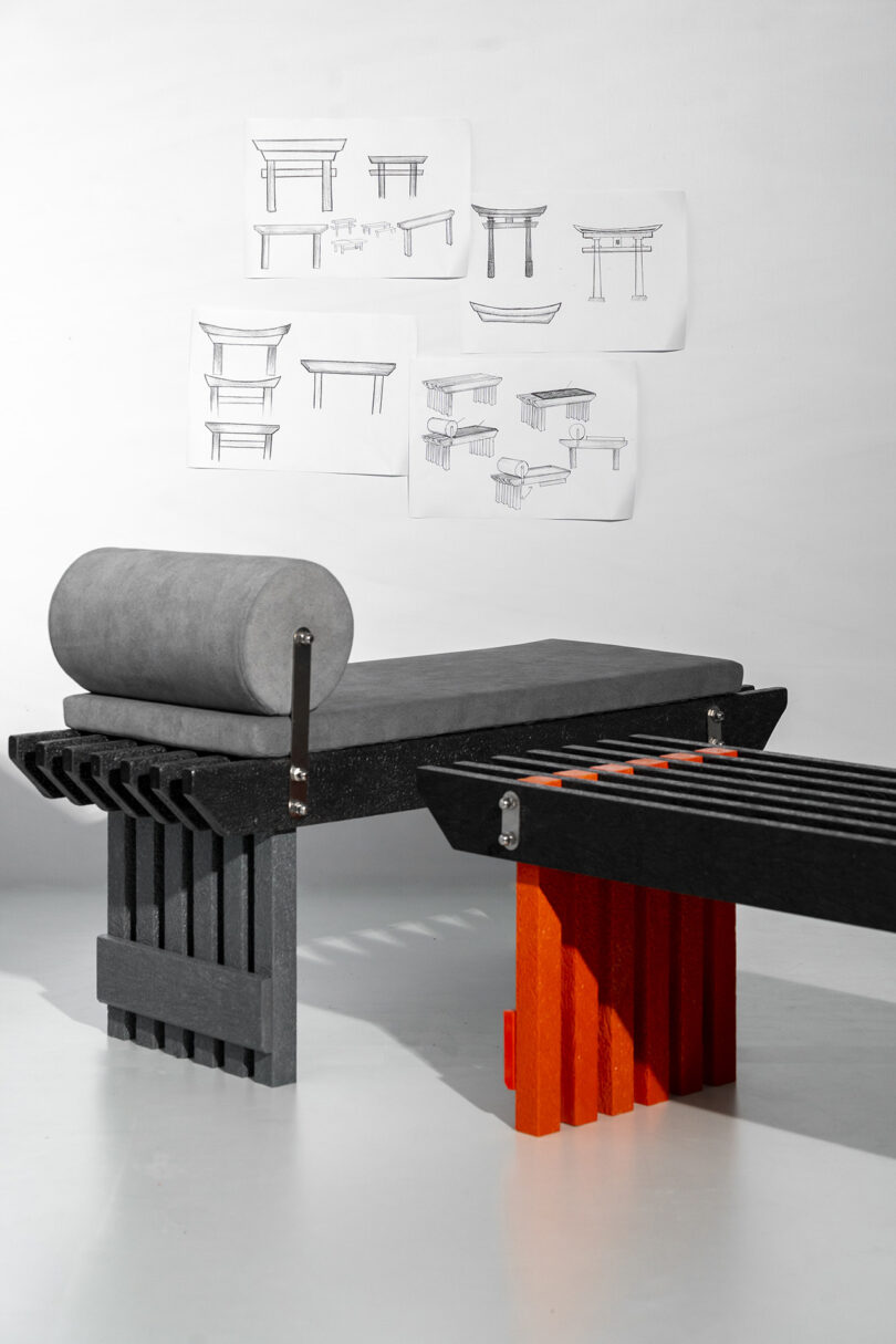 A modern bench with a gray cylindrical cushion and black and red slatted base, positioned in front of sketches of similar furniture designs taped to a white wall.