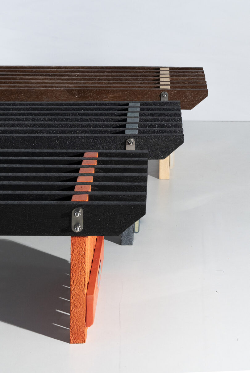 Three modern benches with slatted wooden tops and colored legs in black, brown, and orange are arranged in parallel on a white floor.