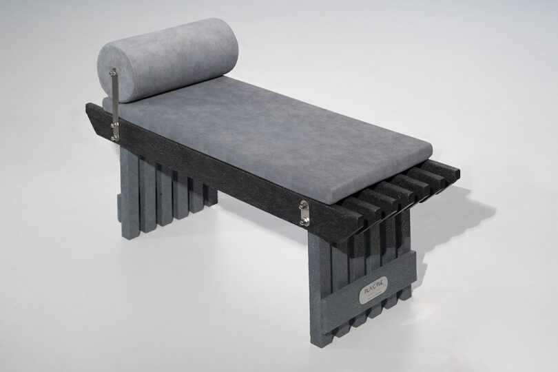 A modern bench with a grey cushioned seat and cylindrical headrest, supported by a black and grey slatted wooden base, against a plain white background.