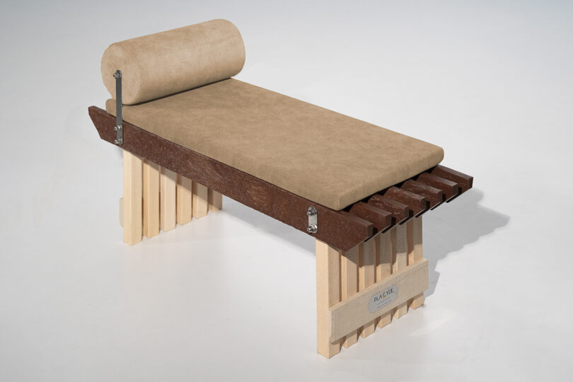 A modern bench with a cushioned beige seat and cylindrical headrest, supported by a structure of wooden slats and metal brackets.