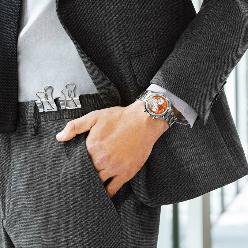 A person in a gray suit with binder clips attached to their waistband, wearing a watch, and resting one hand in their pocket.