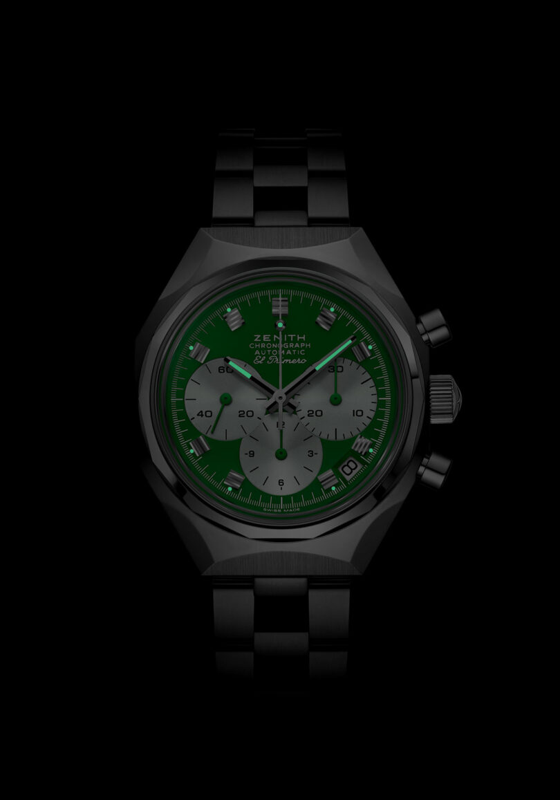 A black metal wristwatch with a green face, silver subdials, and three side buttons is displayed against a black background.