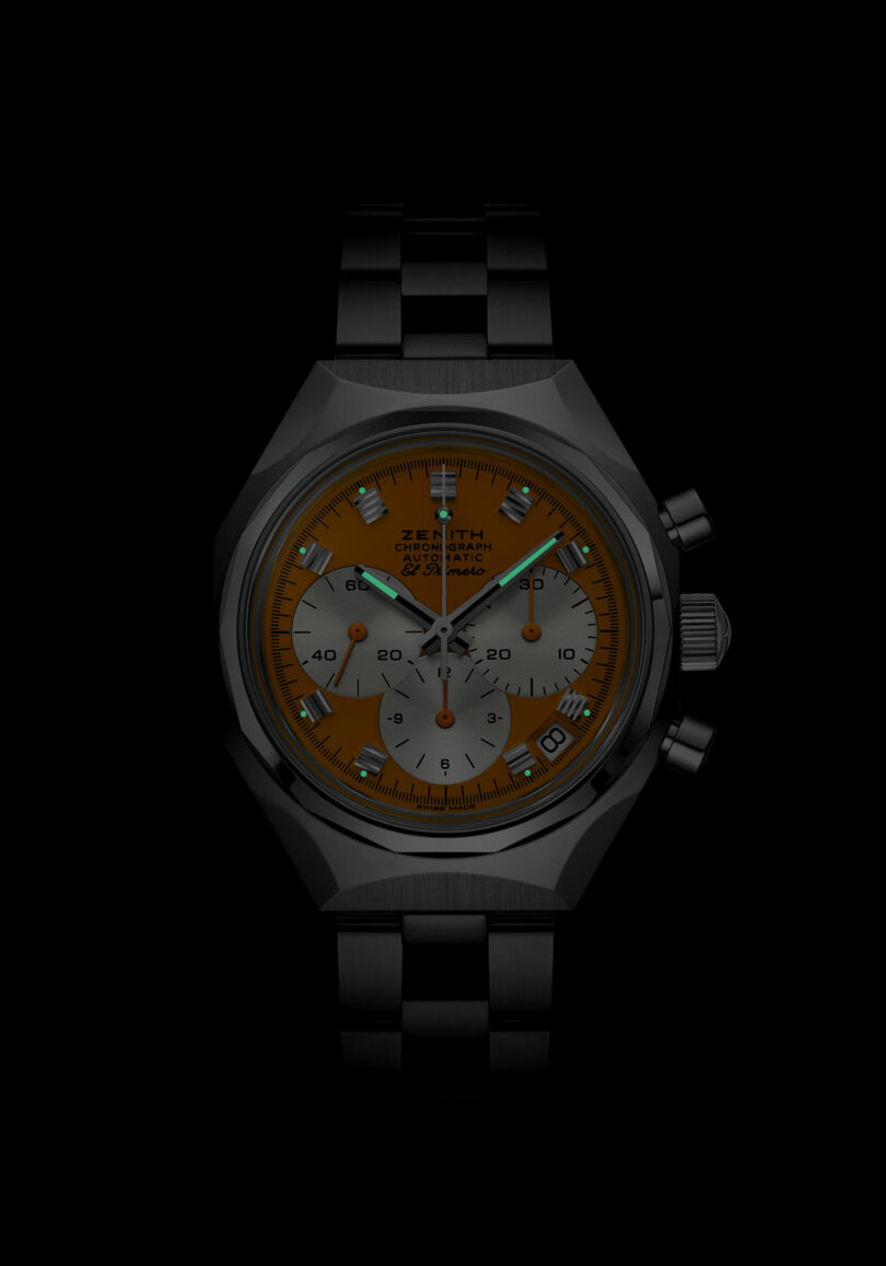 A wristwatch with a black metal band and case, featuring an orange dial and three subdials, with luminous hands and markers, photographed against a black background.