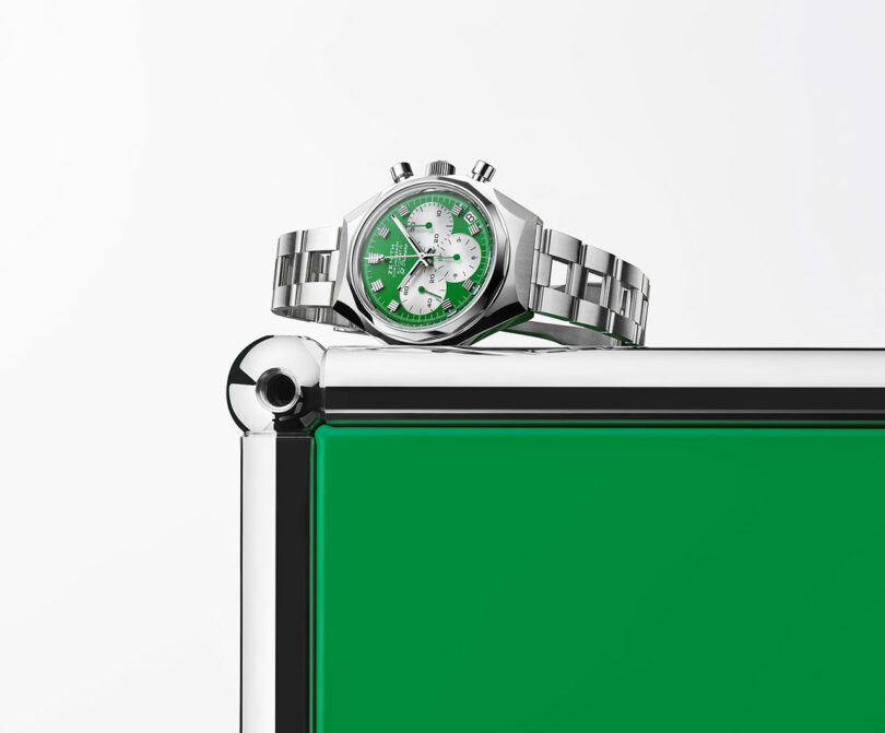 A silver wristwatch with a green and white face rests on the corner of a metallic and green rectangular structure against a plain background.