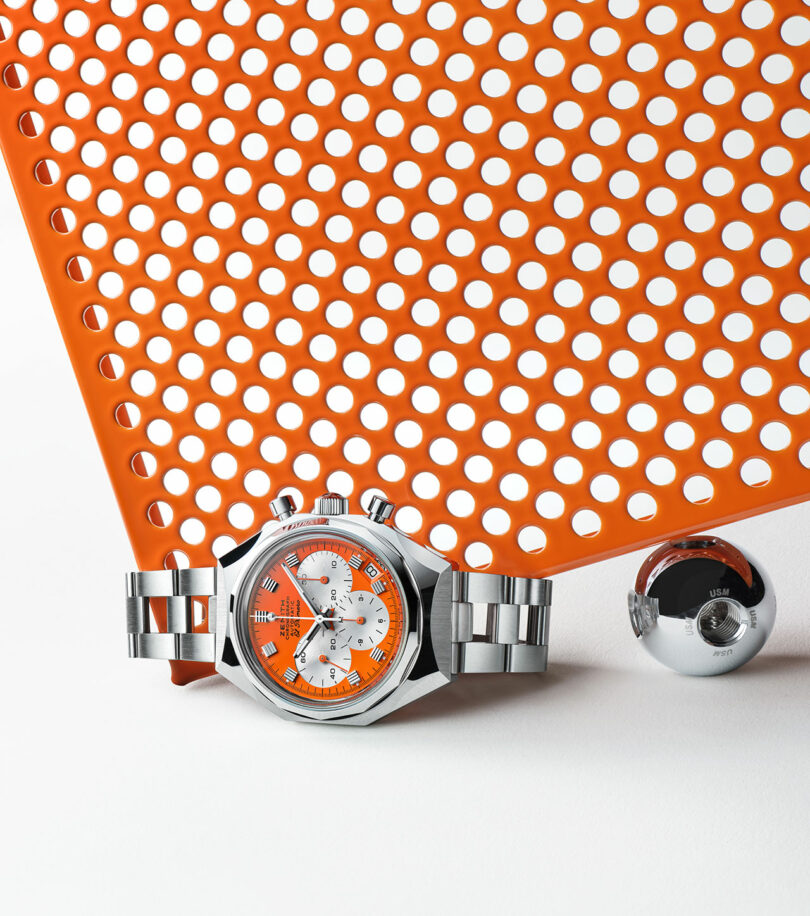 A stainless steel wristwatch with an orange and white face is displayed on a white surface beneath an orange perforated sheet and near a small metallic sphere.