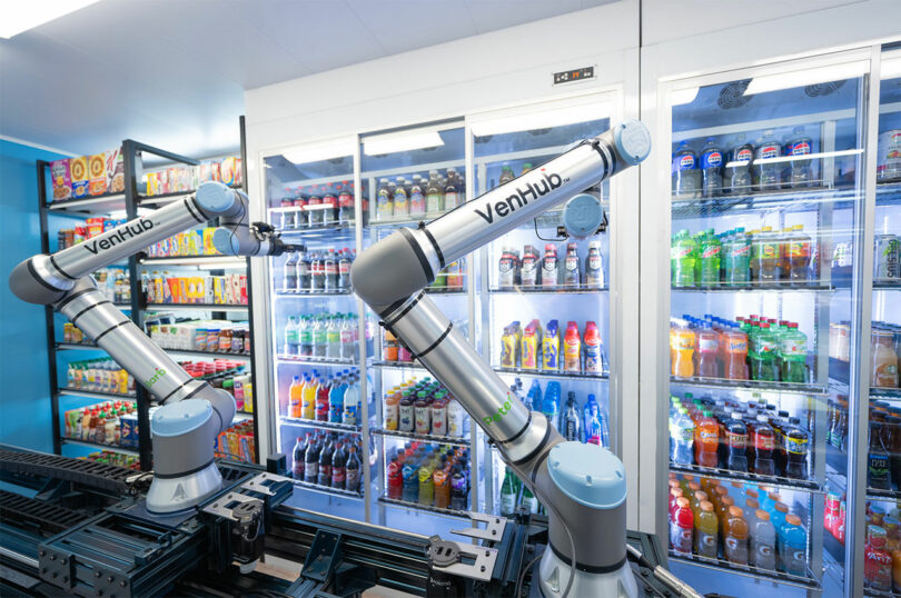 Two robotic arms labeled "VenHub" operate in front of refrigerated shelves stocked with various drinks and food products in a modern automated store.