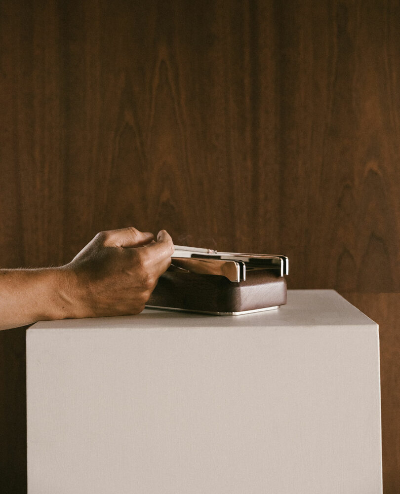 A hand rests on a rectangular wooden object with a metal handle, placed on a white pedestal against a wood-paneled background.
