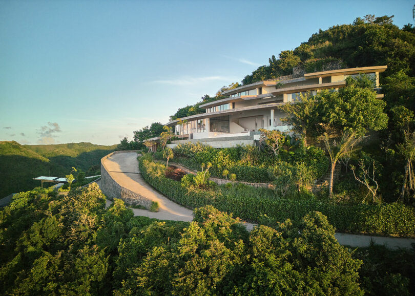 Modern house situated on a hillside surrounded by lush greenery, with a curved driveway leading up to the residence under a clear sky.