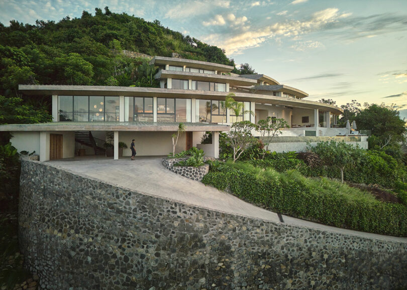 Modern multi-level house built into a hillside, featuring large glass windows, surrounded by greenery and stone retaining walls. A person is walking on the driveway.