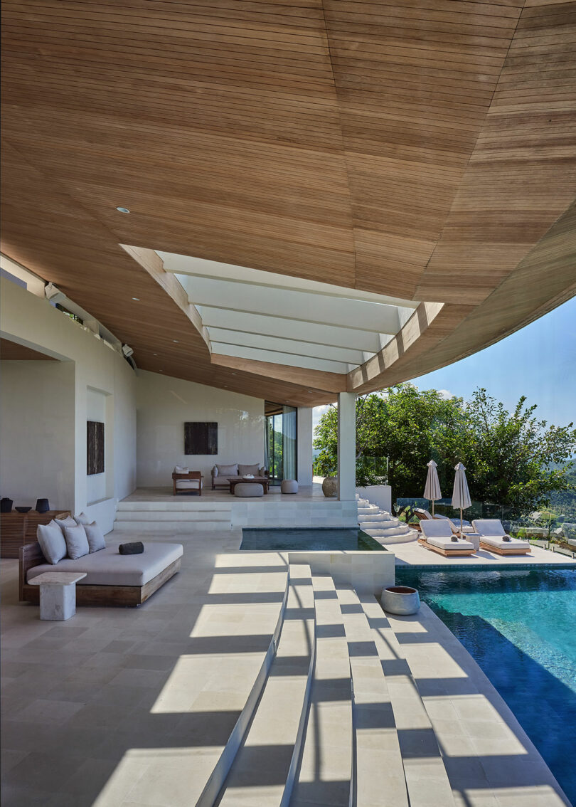 Modern outdoor patio with wooden ceiling, white stone steps, lounge chairs, and a swimming pool, overlooking greenery and sky in the background.