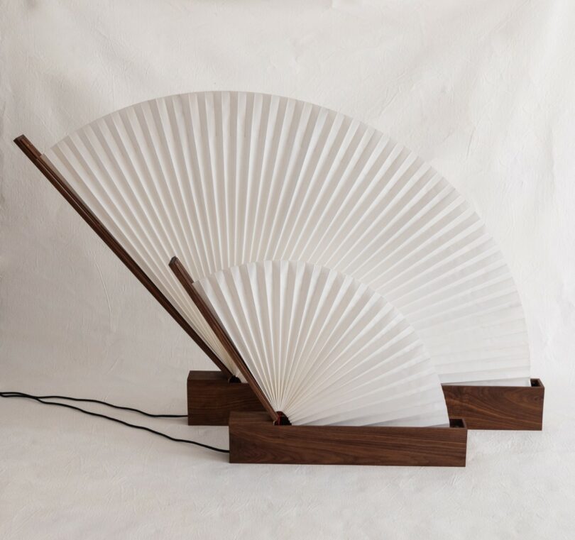 two lamps with a pleated, fan-shaped white shade and a wooden base, connected to a black power cord, set against a plain background