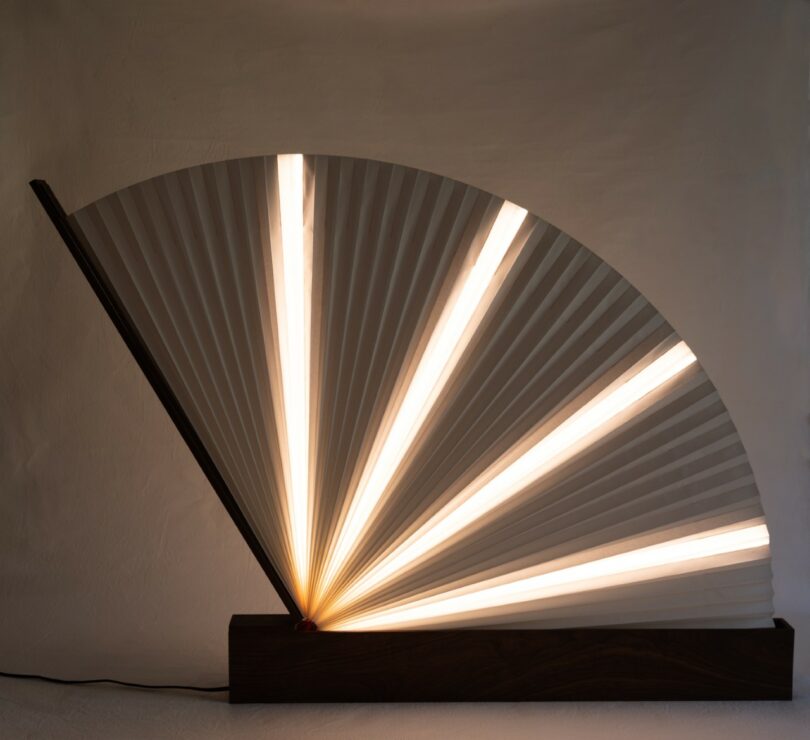 A lamp with a wooden rectangular base and a fan-shaped paper shade emits warm light, positioned against a plain, textured wall