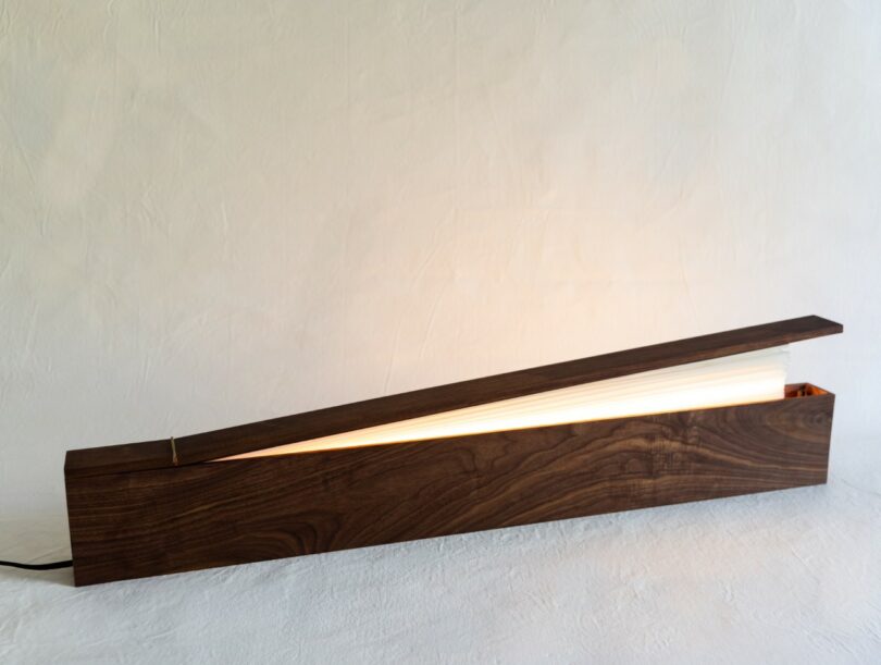 A modern wooden desk lamp with an angled rectangular base and an illuminated strip beneath a partially open wooden lid