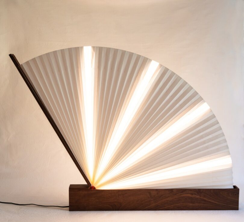 A lamp with a wooden rectangular base and a fan-shaped paper shade emits warm light, positioned against a plain, textured wall