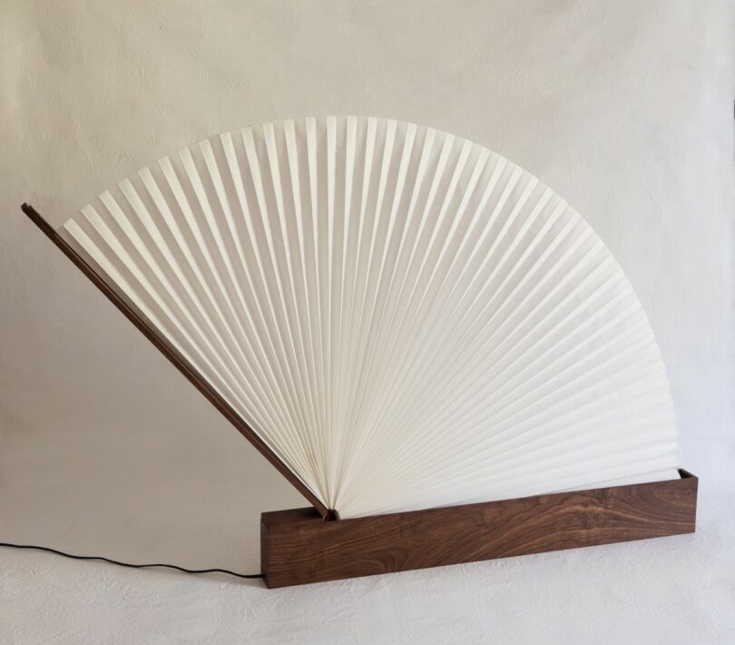 A lamp with a pleated, fan-shaped white shade and a wooden base, connected to a black power cord, set against a plain background