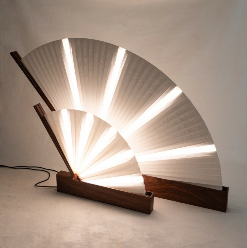 A modern lamp with two fan-shaped white shades and wooden bases, illuminated with vertical LED light strips, placed against a neutral background