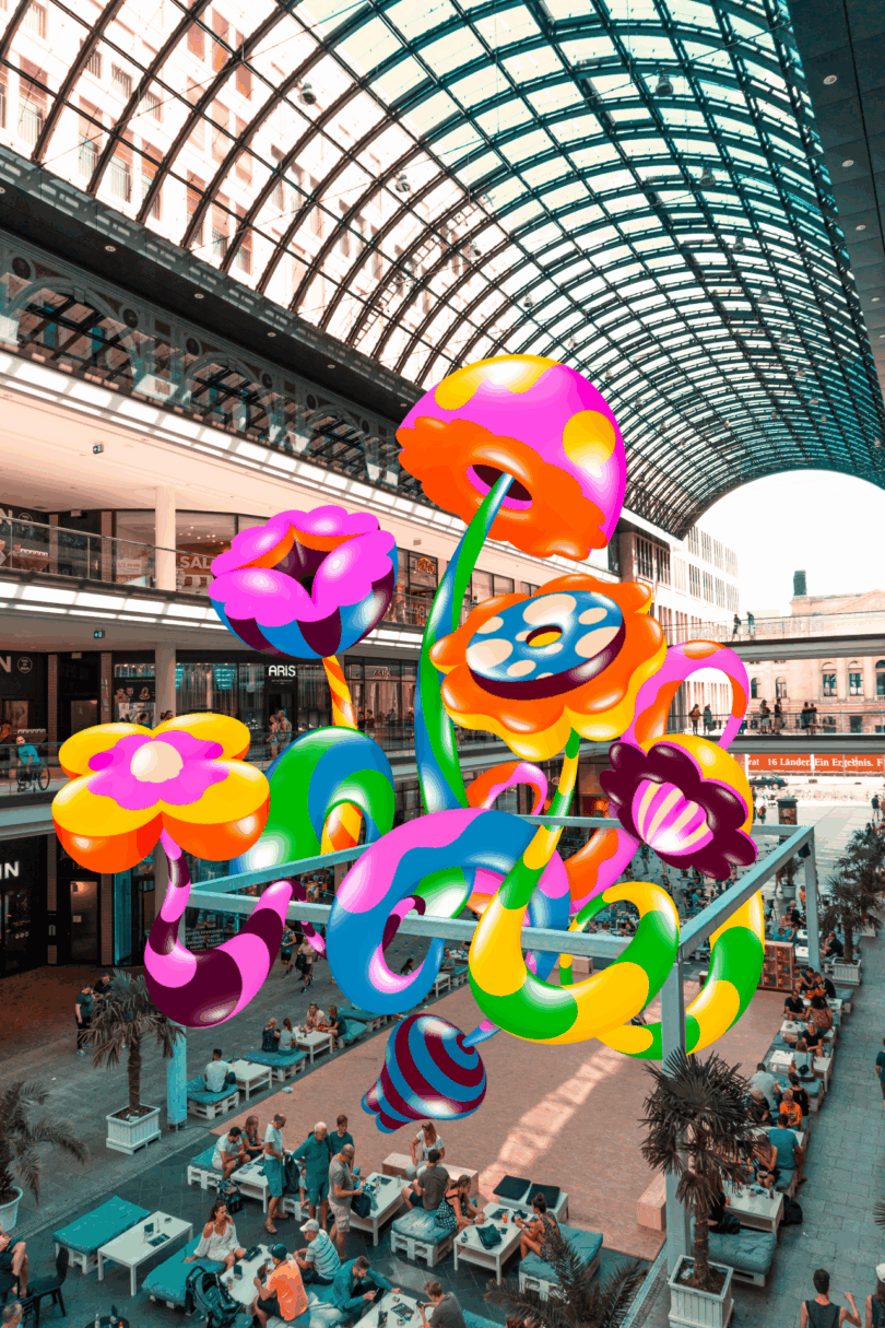 A large, colorful, cartoon-style flower sculpture is suspended above an indoor mall's dining area with people seated below and a glass arched ceiling overhead