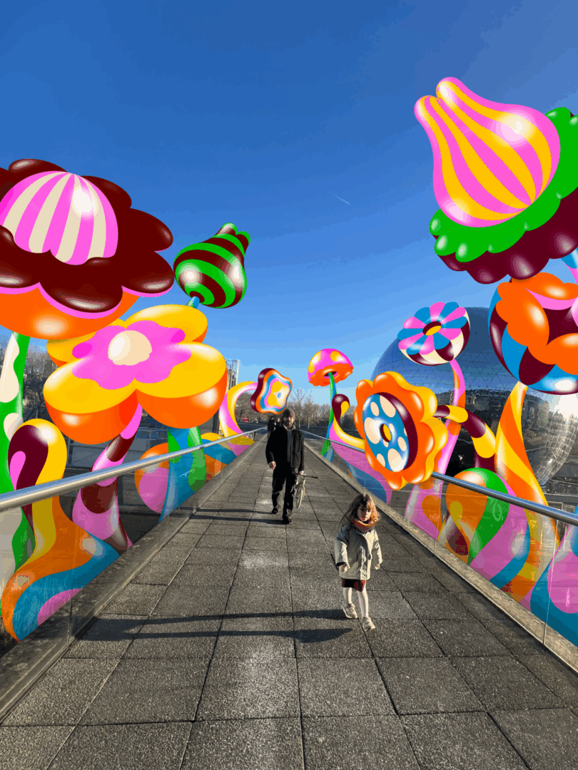 A man and a girl walk on a pedestrian bridge surrounded by large, colorful, cartoon-like inflatable flowers under a clear blue sky